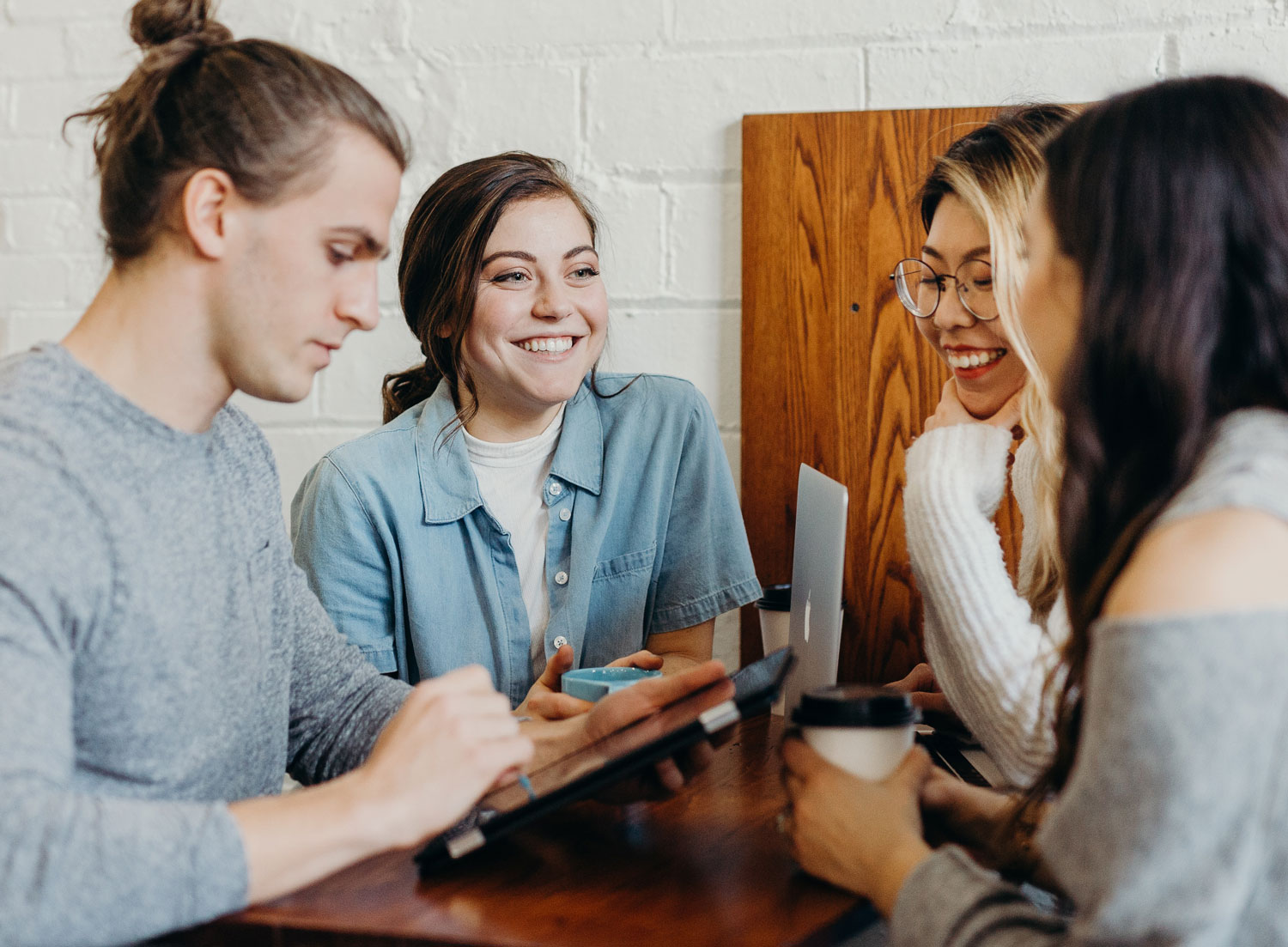 Vier junge Menschen mit Kaffeebechern und Laptops sitzen  am Tisch und unterhalten sich lachend 